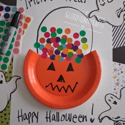 A pumpkin-themed Halloween craft made with an orange paper plate, colorful dot stickers, and ghost drawings on white paper that says Trick or Treat and Happy Halloween!.