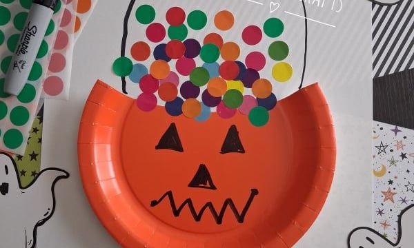 A pumpkin-themed Halloween craft made with an orange paper plate, colorful dot stickers, and ghost drawings on white paper that says Trick or Treat and Happy Halloween!.