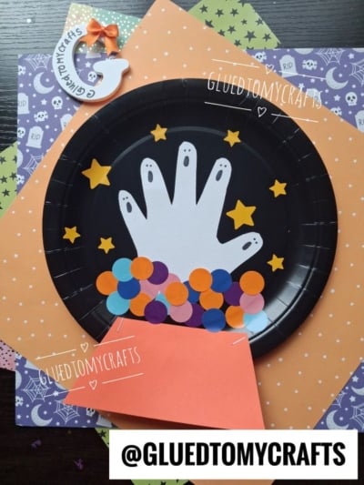 A black plate with a white paper hand, multicolored paper dots, and yellow stars makes a playful scene, perfect for Halloween Crafts for Kids. An orange paper cone shapes the bottom, while Halloween-themed paper is used as a background. @GLUEDTOMYCRAFTS is written on the image.