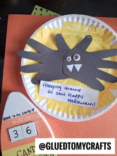 A paper plate Halloween craft featuring a black spider with googly eyes and fangs, yellow tissue background, and a note saying, Hanging around to say Happy Halloween! Labeled @GLUEDTOMYCRAFTS.