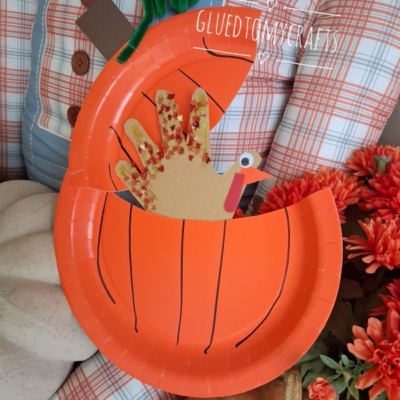 Paper pumpkin plate craft with a handprint turkey peeking out, set among fall decor including a scarecrow and orange flowers. The image is watermarked @GLUEDTOMYCRAFTS.