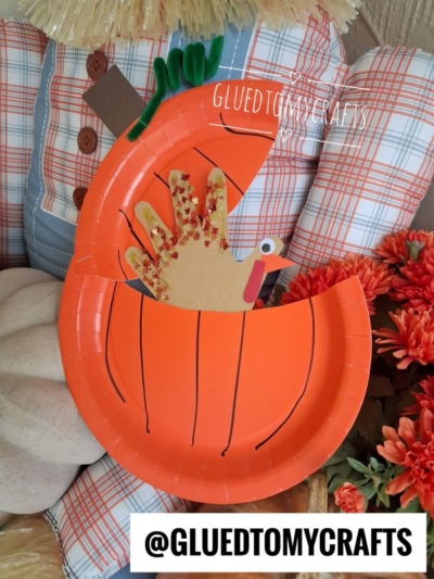 Paper pumpkin plate craft with a handprint turkey peeking out, set among fall decor including a scarecrow and orange flowers. The image is watermarked @GLUEDTOMYCRAFTS.