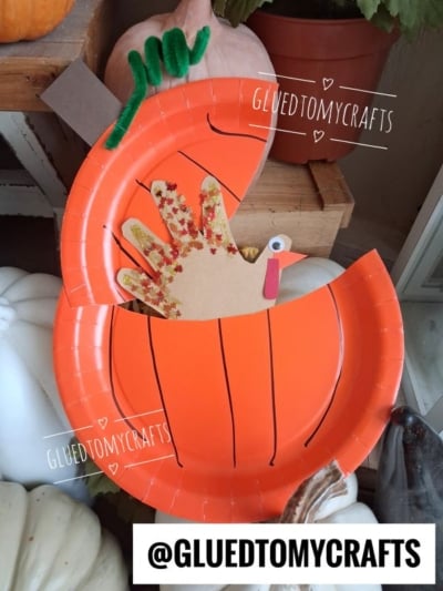 A paper plate pumpkin opens to reveal a hiding turkey craft made from construction paper and decorated with glitter. The craft sits among white pumpkins and fall decor.