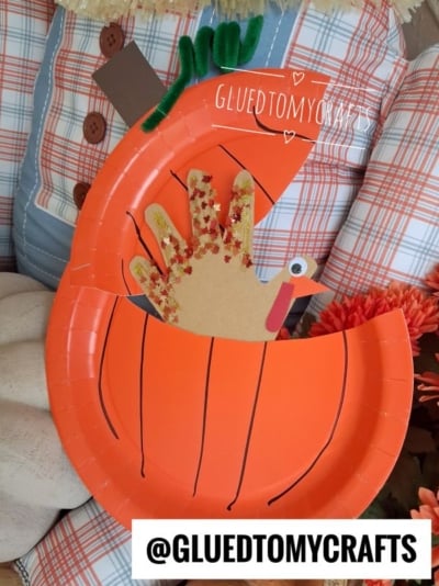 A paper plate pumpkin craft with a handprint turkey inside, decorated with glitter and googly eyes, set against plaid fabric and pumpkins. The handle @GLUEDTOMYCRAFTS is written on the image.