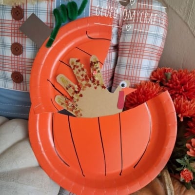 Paper plate pumpkin craft with a handprint turkey inside. The pumpkin is orange with drawn lines, and the turkey has a googly eye and red glitter on its handprint feathers. Text reads @GLUEDTOMYCRAFTS.