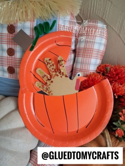Paper plate pumpkin craft with a handprint turkey inside. The pumpkin is orange with drawn lines, and the turkey has a googly eye and red glitter on its handprint feathers. Text reads @GLUEDTOMYCRAFTS.