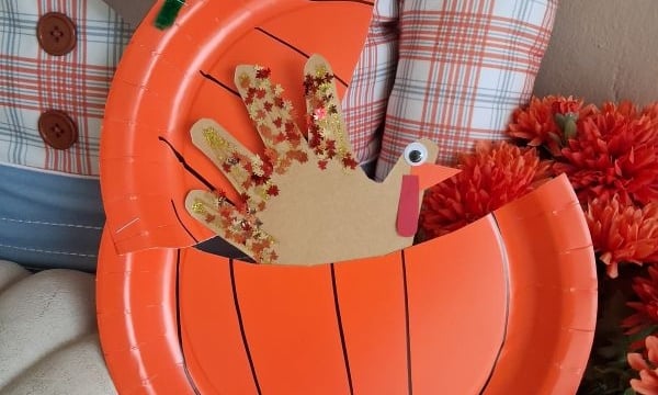 Paper plate pumpkin craft with a handprint turkey inside. The pumpkin is orange with drawn lines, and the turkey has a googly eye and red glitter on its handprint feathers. Text reads @GLUEDTOMYCRAFTS.