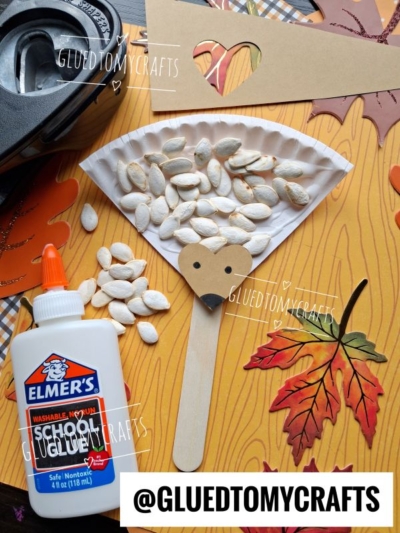 A paper plate hedgehog craft with pumpkin seeds, glue, brown heart face, and craft stick body is displayed on a fall-themed surface with leaves and tape in the background.