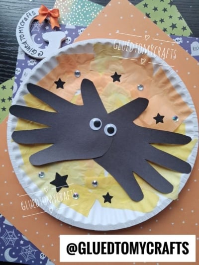 A paper plate craft with orange, yellow, and white tissue paper, two black paper handprints for a spider, googly eyes, black stars, and gems. Halloween-themed paper is in the background.