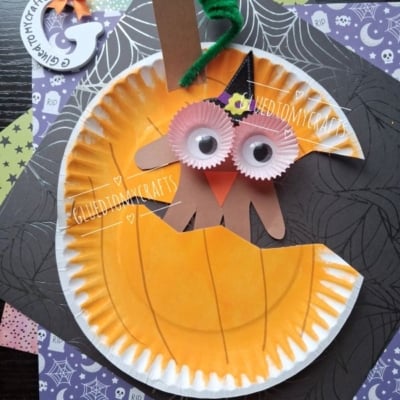 Paper plate pumpkin craft with googly eyes, orange cupcake liners, a green pipe cleaner stem, and a brown paper mouth, set on Halloween-themed paper. @GLUEDTOMYCRAFTS is written at the bottom.