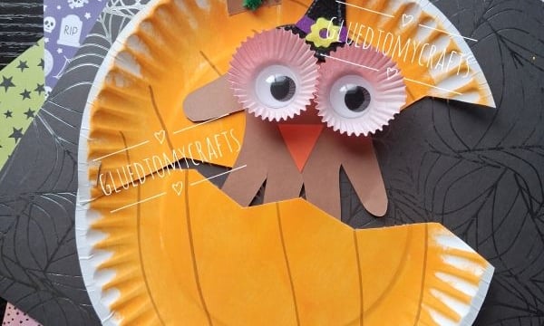 Paper plate pumpkin craft with googly eyes, orange cupcake liners, a green pipe cleaner stem, and a brown paper mouth, set on Halloween-themed paper. @GLUEDTOMYCRAFTS is written at the bottom.