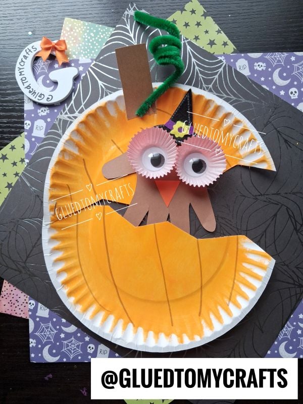 Paper plate pumpkin craft with googly eyes, orange cupcake liners, a green pipe cleaner stem, and a brown paper mouth, set on Halloween-themed paper. @GLUEDTOMYCRAFTS is written at the bottom.