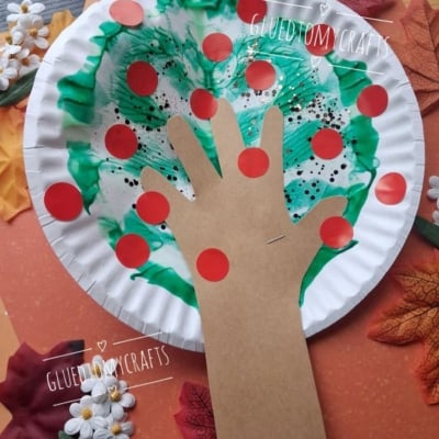A paper plate craft of an apple tree with a brown paper hand as the trunk, green and white paint for leaves, red dots as apples, and fake leaves and flowers around it. @GLUEDTOMYCRAFTS is written on the image.