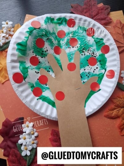 A paper plate craft of an apple tree with a brown paper hand as the trunk, green and white paint for leaves, red dots as apples, and fake leaves and flowers around it. @GLUEDTOMYCRAFTS is written on the image.