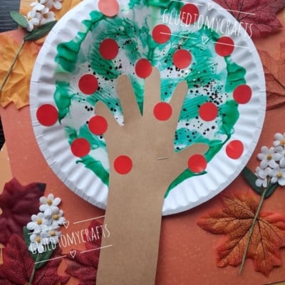 Paper plate craft of a hand-shaped brown paper cutout as a tree trunk, green paint as leaves, and red dots as apples. Surrounded by autumn leaves, flowers, and orange paper. Text: @GLUEDTOMYCRAFTS.