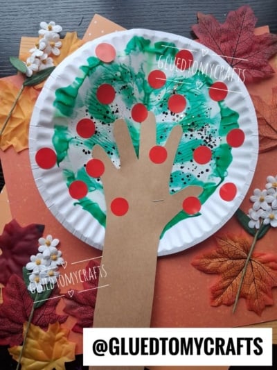 Paper plate craft of a hand-shaped brown paper cutout as a tree trunk, green paint as leaves, and red dots as apples. Surrounded by autumn leaves, flowers, and orange paper. Text: @GLUEDTOMYCRAFTS.
