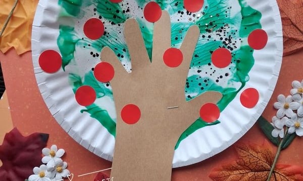 Paper plate craft of a hand-shaped brown paper cutout as a tree trunk, green paint as leaves, and red dots as apples. Surrounded by autumn leaves, flowers, and orange paper. Text: @GLUEDTOMYCRAFTS.