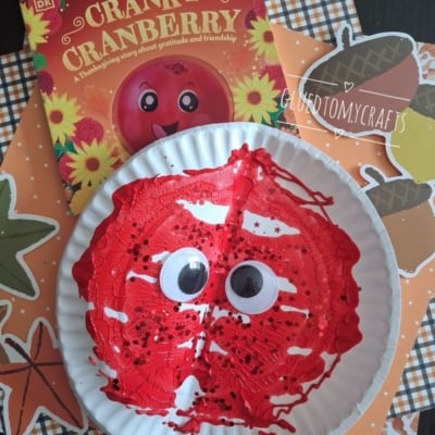 A paper plate painted red with googly eyes resembles a cranberry, placed on fall-themed paper next to the book The Cranky Cranberry. Text @GLUEDTOMYCRAFTS is at the bottom.