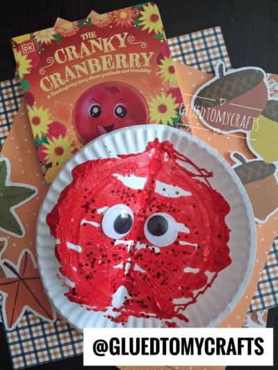 A paper plate painted red with googly eyes resembles a cranberry, placed on fall-themed paper next to the book The Cranky Cranberry. Text @GLUEDTOMYCRAFTS is at the bottom.