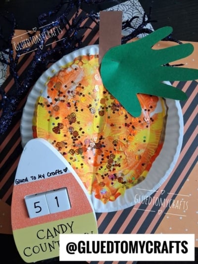 A pumpkin craft made from a paper plate with orange paint, sequins, and a green handprint for a leaf sits on a Halloween-themed background. Perfect for Halloween Crafts for Kids, this project also features CANDY COUNTDOWN 51 and @GLUEDTOMYCRAFTS in the scene.