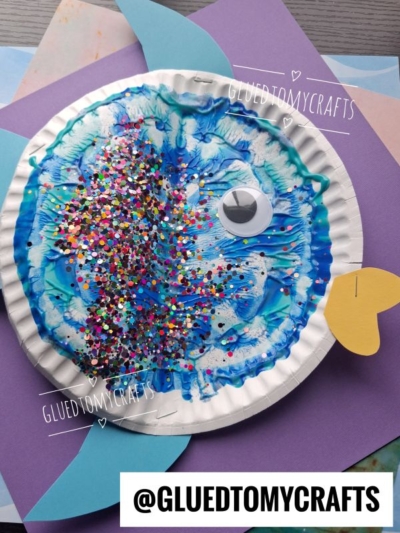 A paper plate fish craft with blue paint, glitter, a large googly eye, blue fin cutouts, and a yellow paper tail. The fish is placed on colorful paper. @GLUEDTOMYCRAFTS is at the bottom.