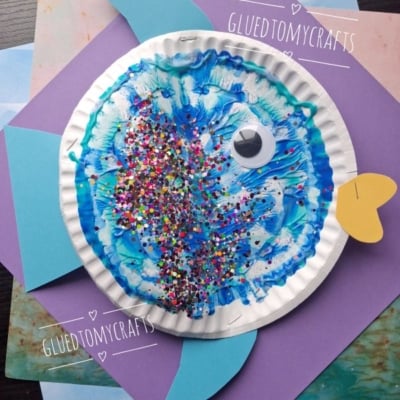 A colorful fish craft made from a paper plate, blue paint, glitter, a googly eye, and colored paper fins sits on layered sheets of purple and blue paper. @GLUEDTOMYCRAFTS is written on the image.