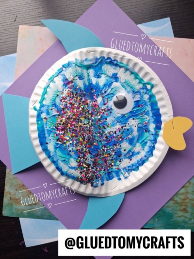 A colorful fish craft made from a paper plate, blue paint, glitter, a googly eye, and colored paper fins sits on layered sheets of purple and blue paper. @GLUEDTOMYCRAFTS is written on the image.