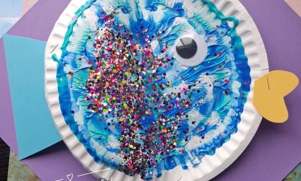 A colorful fish craft made from a paper plate, blue paint, glitter, a googly eye, and colored paper fins sits on layered sheets of purple and blue paper. @GLUEDTOMYCRAFTS is written on the image.
