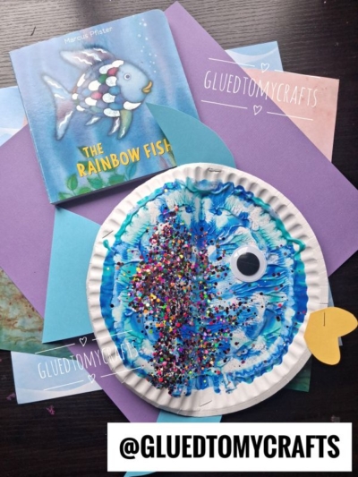 A Rainbow Fish craft with a paper plate, paint, glitter, and a googly eye sits on colored paper next to the book The Rainbow Fish by Marcus Pfister. The username @GLUEDTOMYCRAFTS is labeled on the image.