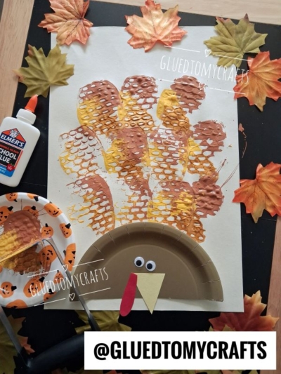 A kids turkey craft made with a brown painted paper plate, googly eyes, a red wattle, a yellow beak, and potato masher stamped craft feathers. Surrounded by fake autumn leaves and crafting materials.