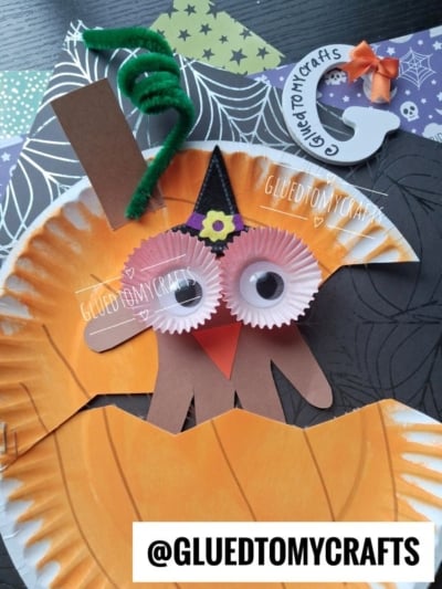 A handmade owl craft using orange paper plate, cupcake liners for eyes, brown paper wings, and a black hat, with @gluedtomycrafts text in two spots and crafting materials in the background.