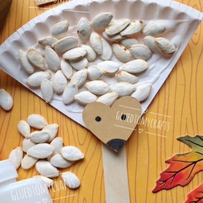 A paper plate hedgehog craft with a popsicle stick handle, pumpkin seeds for spikes, and a paper face, surrounded by craft supplies and fall leaves.