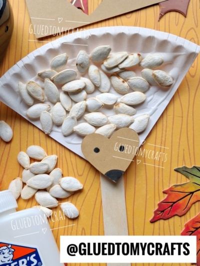 A paper plate hedgehog craft with a popsicle stick handle, pumpkin seeds for spikes, and a paper face, surrounded by craft supplies and fall leaves.