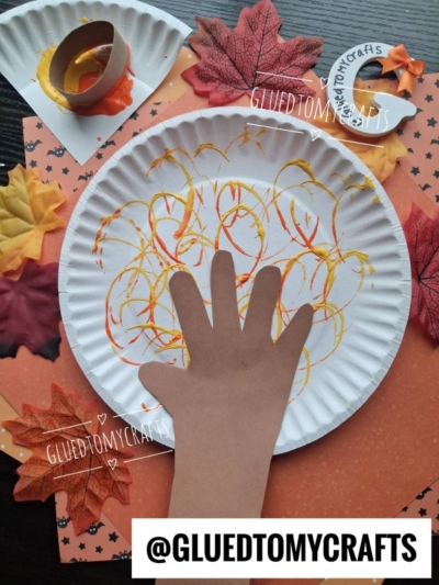 A paper craft campfire with a brown handprint cutout as the log and painted orange, red, and yellow flames on a paper plate, surrounded by artificial autumn leaves—this is a perfect example of fun FALL CRAFTS FOR KIDS.