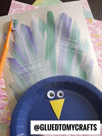 A peacock craft made with a blue paper plate, googly eyes, a yellow beak, and painted feather strokes in green and purple. A toothbrush is used for painting. @gluedtomycrafts is prominently displayed.