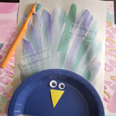 A kids craft of a peacock with a blue paper plate body, googly eyes, a yellow paper beak, and painted feather lines made with a toothbrush. Various pastel papers are in the background. @GLUEDTOMYCRAFTS text is visible.
