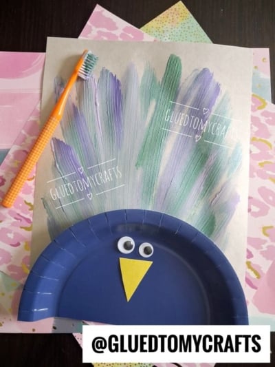 A kids craft of a peacock with a blue paper plate body, googly eyes, a yellow paper beak, and painted feather lines made with a toothbrush. Various pastel papers are in the background. @GLUEDTOMYCRAFTS text is visible.