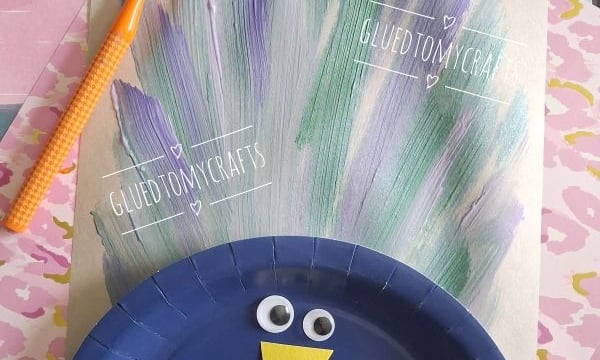 A kids craft of a peacock with a blue paper plate body, googly eyes, a yellow paper beak, and painted feather lines made with a toothbrush. Various pastel papers are in the background. @GLUEDTOMYCRAFTS text is visible.
