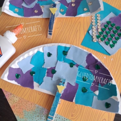 Two paper plate fans decorated with blue and purple paper squares, green heart stickers, rhinestones, and clothespins, displayed on a wooden-pattern background with craft supplies visible.
