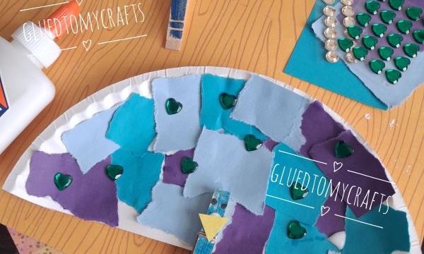 Two paper plate fans decorated with blue and purple paper squares, green heart stickers, rhinestones, and clothespins, displayed on a wooden-pattern background with craft supplies visible.