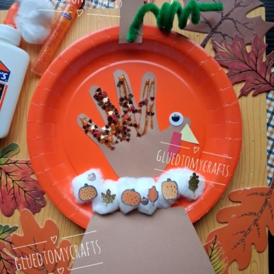 A Thanksgiving turkey craft made from an orange paper plate, brown paper hand cutout, decorated with glitter, pom-poms, mini pumpkin stickers, cotton balls, and green pipe cleaner, placed on a leaf-patterned mat.