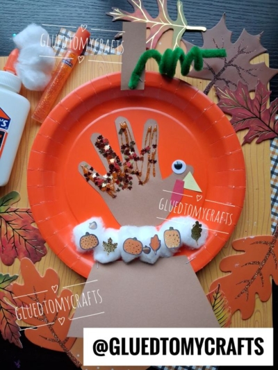 A Thanksgiving turkey craft made from an orange paper plate, brown paper hand cutout, decorated with glitter, pom-poms, mini pumpkin stickers, cotton balls, and green pipe cleaner, placed on a leaf-patterned mat.
