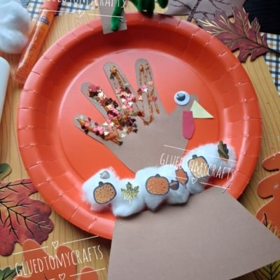 A turkey craft made from a hand-shaped cutout on an orange paper plate, decorated with glitter, pom-poms, paper shapes, and stickers, surrounded by fall leaves. A glue stick and pipe cleaner are nearby.