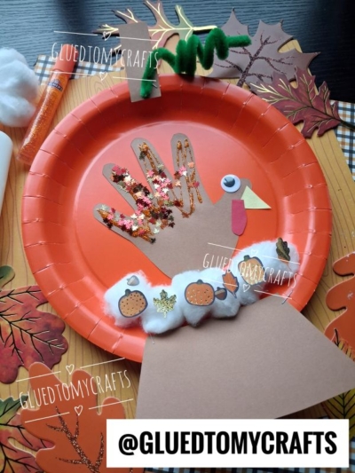 A turkey craft made from a hand-shaped cutout on an orange paper plate, decorated with glitter, pom-poms, paper shapes, and stickers, surrounded by fall leaves. A glue stick and pipe cleaner are nearby.