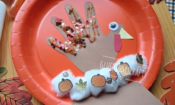 A turkey craft made from a hand-shaped cutout on an orange paper plate, decorated with glitter, pom-poms, paper shapes, and stickers, surrounded by fall leaves. A glue stick and pipe cleaner are nearby.