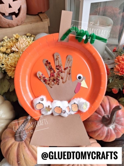 An orange paper plate turkey snow globe craft with a handprint body, glitter, cotton, and paper decorations. Surrounded by artificial fall flowers and pumpkins. @GLUEDTOMYCRAFTS watermark is present.