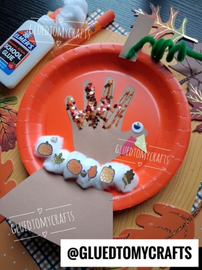 A Thanksgiving turkey craft on an orange plate, made from a handprint, pom-poms, pipe cleaners, foam leaves, and glue. A bottle of Elmers School Glue and fall leaves are in the background. @GLUEDTOMYCRAFTS watermark appears.