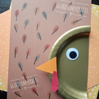 A turkey craft made from brown paper, a gold paper plate, a googly eye, orange beak, and red wattle, with @GLUETOMYCRAFTS text labels and a star-patterned background.