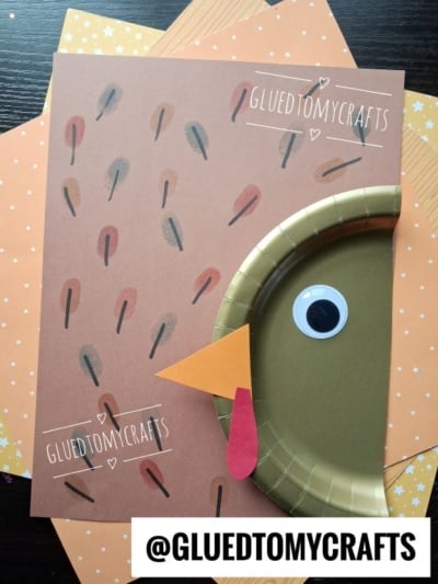 A turkey craft made from brown paper, a gold paper plate, a googly eye, orange beak, and red wattle, with @GLUETOMYCRAFTS text labels and a star-patterned background.