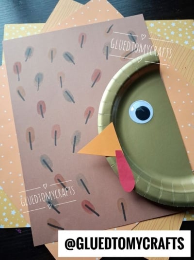 A turkey craft made with a brown paper plate, a googly eye, orange beak, and paper feathers on spotted orange paper. Text reads @GLUEDTOMYCRAFTS and GLUEDTOMYCRAFTS..